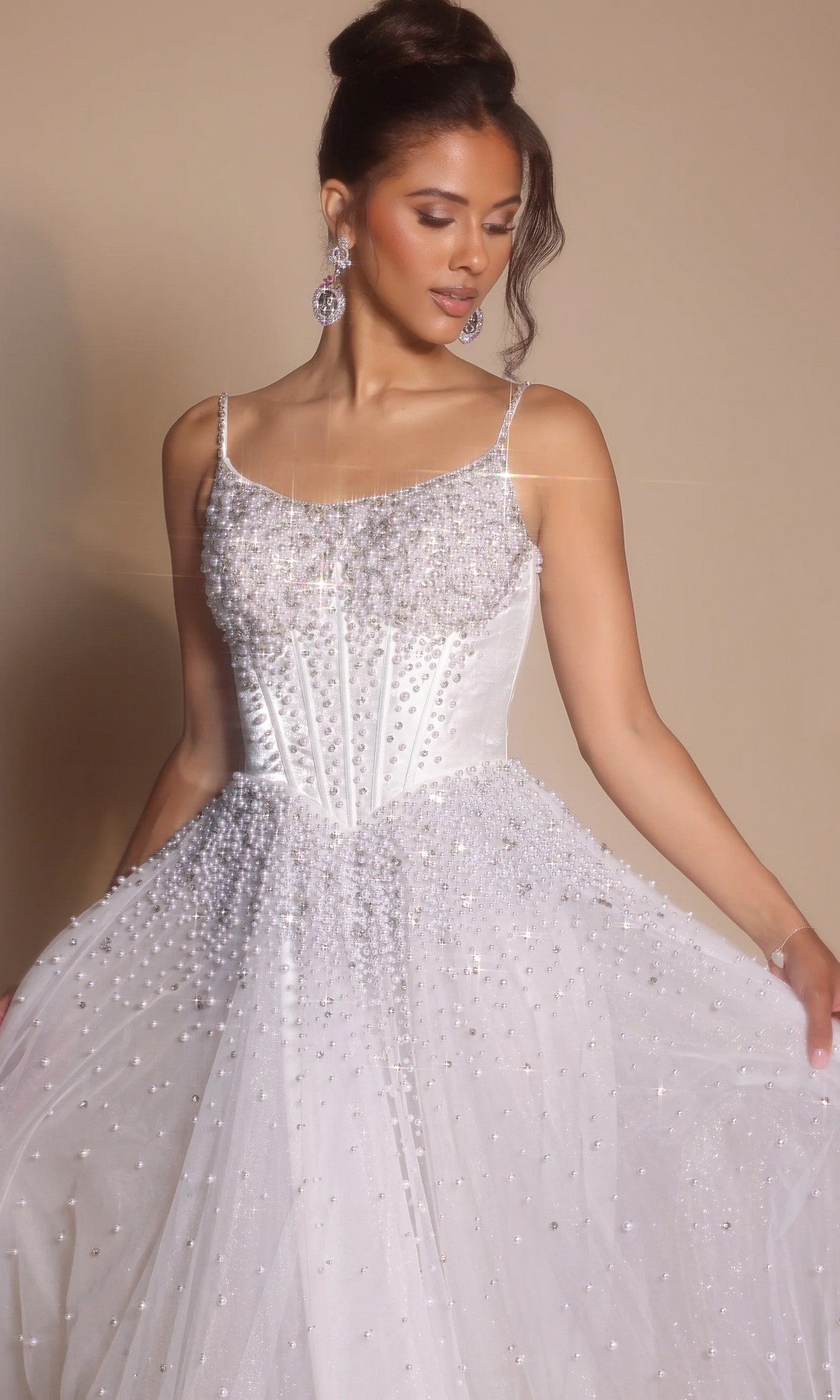 A woman models the Portia and Scarlett PS26208 Corseted Ball Gown in ivory, featuring a beaded boned-corset bodice and thin straps, as she looks down and holds out the skirt against a plain background.